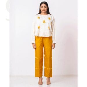 A woman wearing a handmade sustainable co-ord set featuring an ivory top with yellow floral motifs and mustard tie-dye pants, paired with neutral heels. The outfit combines traditional craftsmanship with eco-friendly fashion.