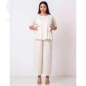 A woman wearing an ivory handmade co-ord set featuring a pleated peplum-style top with side-button details and wide-leg trousers. The outfit exudes minimalist elegance and sustainable fashion.