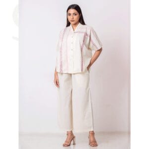 A model wearing a Taan cream-colored short-sleeved button-up shirt with subtle pink vertical patterns, paired with wide-leg cream pants, standing against a plain light background.