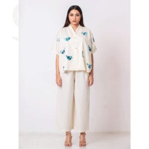 A model wearing a cream-colored short-sleeved button-up shirt adorned with blue bird motifs, paired with wide-leg cream pants, standing against a plain light background.