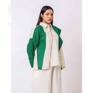 A model wearing a dual-tone button-up shirt featuring green and cream panels with vertical stripes, accented by red-stitched details on the placket, paired with wide-leg cream pants, standing against a plain light background.