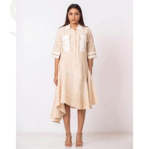 A woman wearing a beige handwoven asymmetrical dress with intricate handcrafted stitch detailing on the sleeves and pockets, styled with a collar and flowy hemline. The dress is paired with neutral heels, showcasing a sustainable and elegant fashion look by Taan.